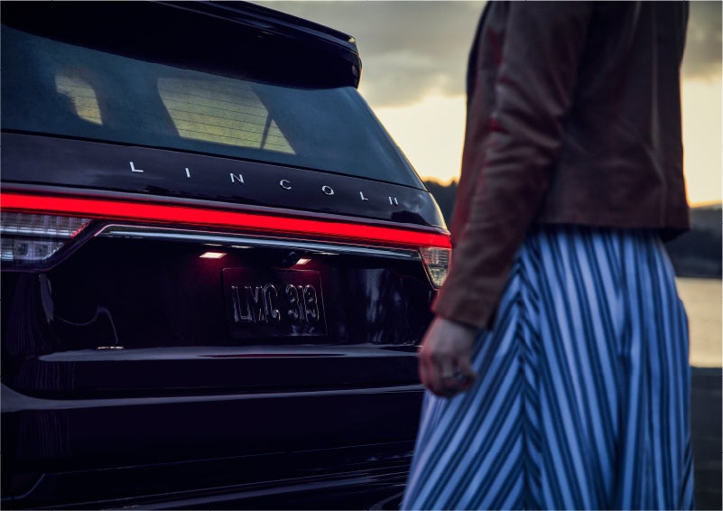 A person is shown near the rear of a 2023 Lincoln Aviator® SUV as the Lincoln Embrace illuminates the rear lights | Evergreen Lincoln in Issaquah WA